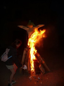 Un pelele y otros enseres arden en la hoguera de San Juan. Una persona echa algo al fuego.